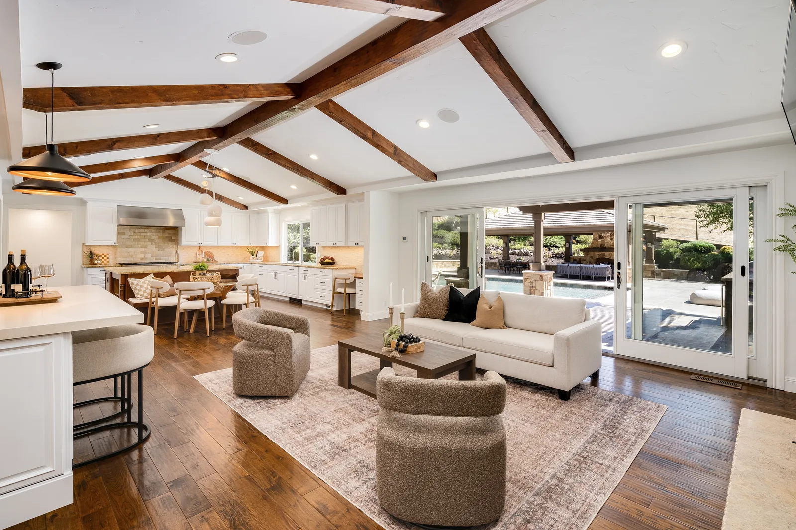 The great room at 2780 Mossy Oak Drive: exposed beams over a vaulted ceiling, seating and bar, with sliders opening onto the pool and stone pavilion