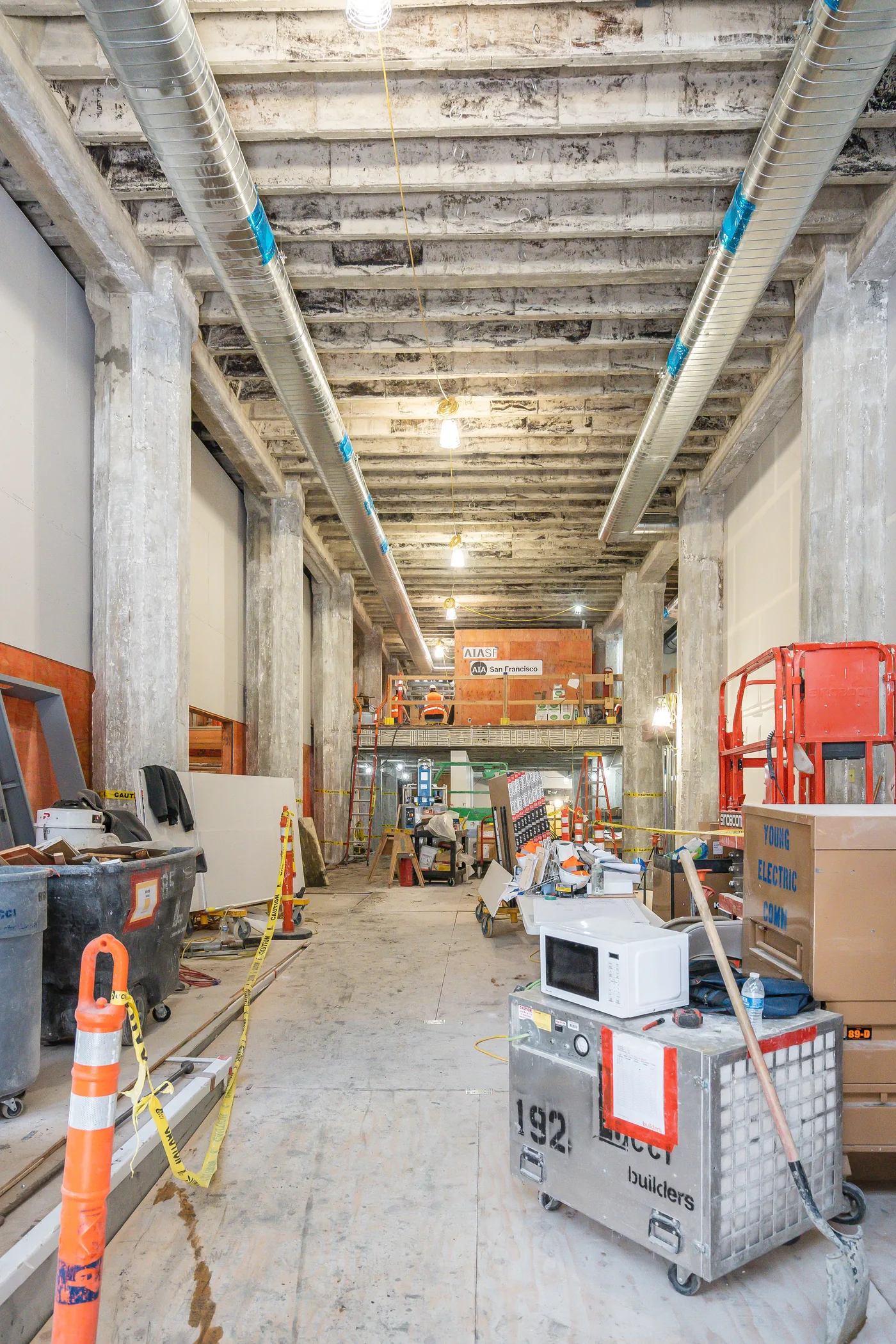 The Center for Architecture + Design mid-construction: exposed concrete beams and ductwork above a gallery of stacked material, scaffolding and site protection, a worker on the mezzanine beneath the AIA San Francisco sign