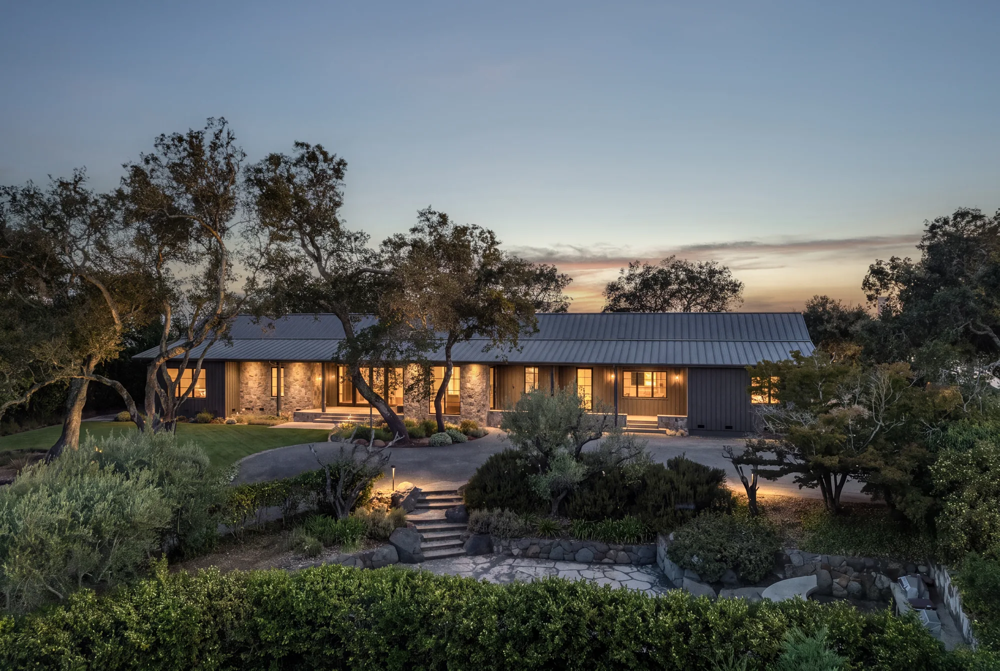 A Napa residence at twilight: a low house under a metal roof among oaks, every window lit warm against a fading sky