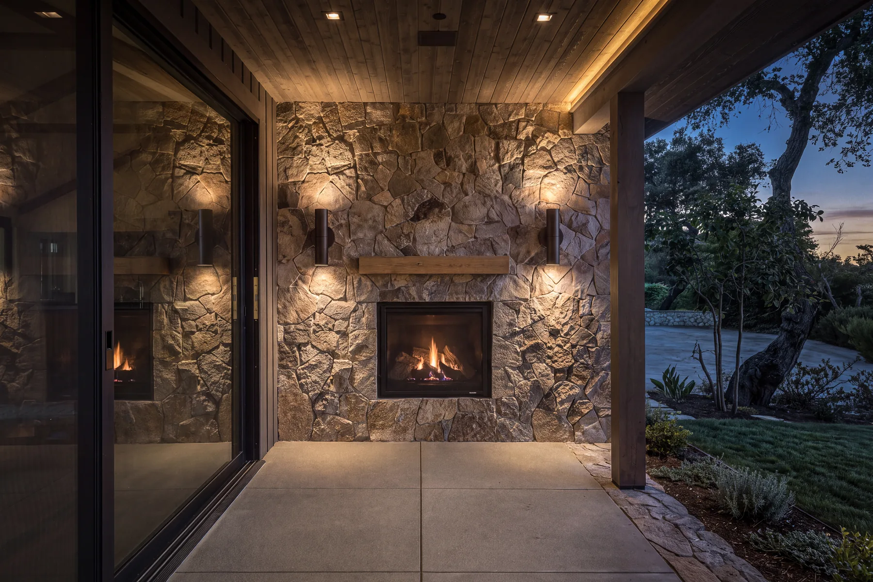 A Napa residence, covered terrace: a lit fireplace set into a stone chimney breast under a timber soffit, sliding doors to one side and oaks beyond