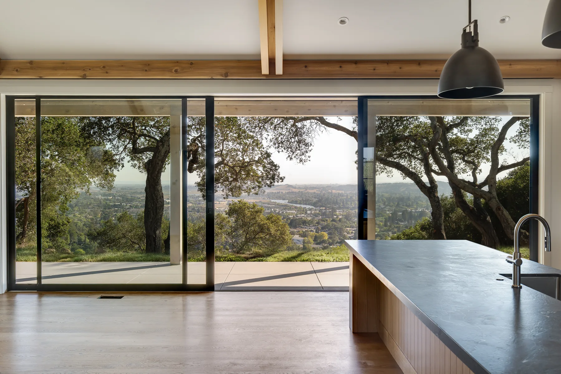 A Napa residence, kitchen: a dark stone island beside sliding doors opened to oaks and the valley beyond