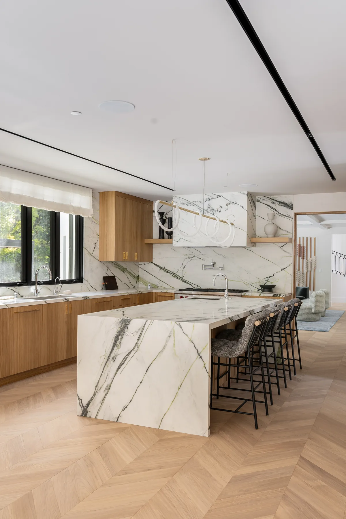 A Silicon Valley residence, kitchen: a marble waterfall island and run of black stools against white oak cabinetry, the veined slab carried up behind the range