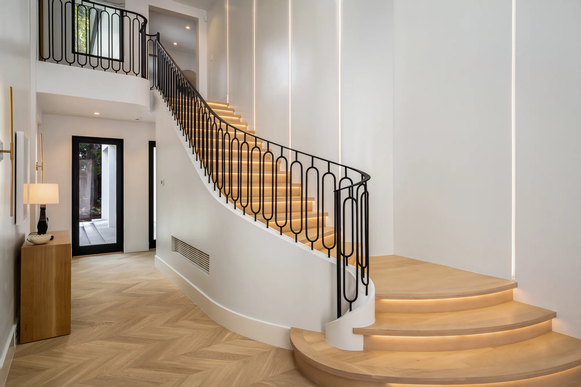 The same entry finished and lit: oak treads glowing from concealed strips, a slim black railing curving down to a herringbone floor, vertical light lines washing the panelled wall