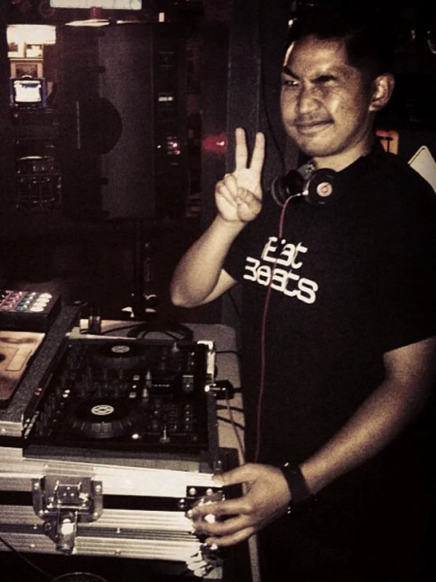 Mike behind a DJ setup, headphones on, throwing a peace sign.